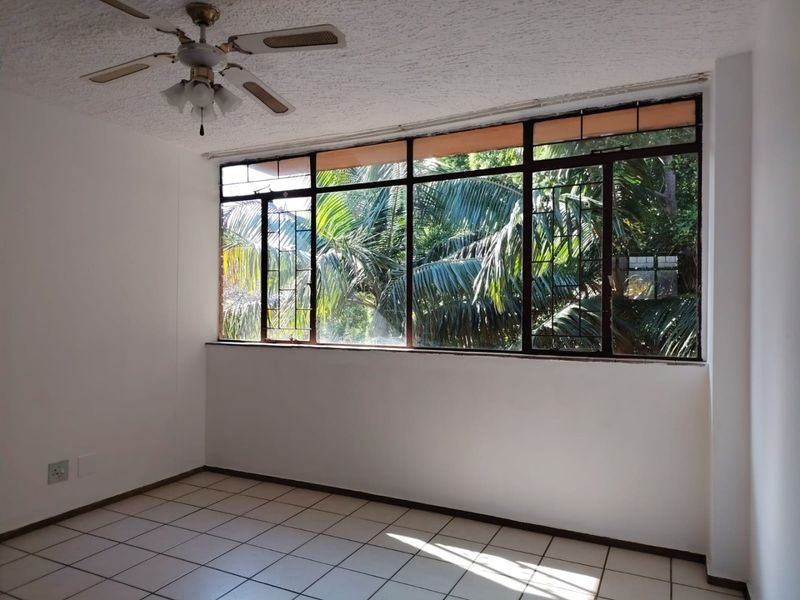 An empty bedroom featuring tiled flooring, a ceiling fan, and large metal-framed windows with a view of lush greenery.