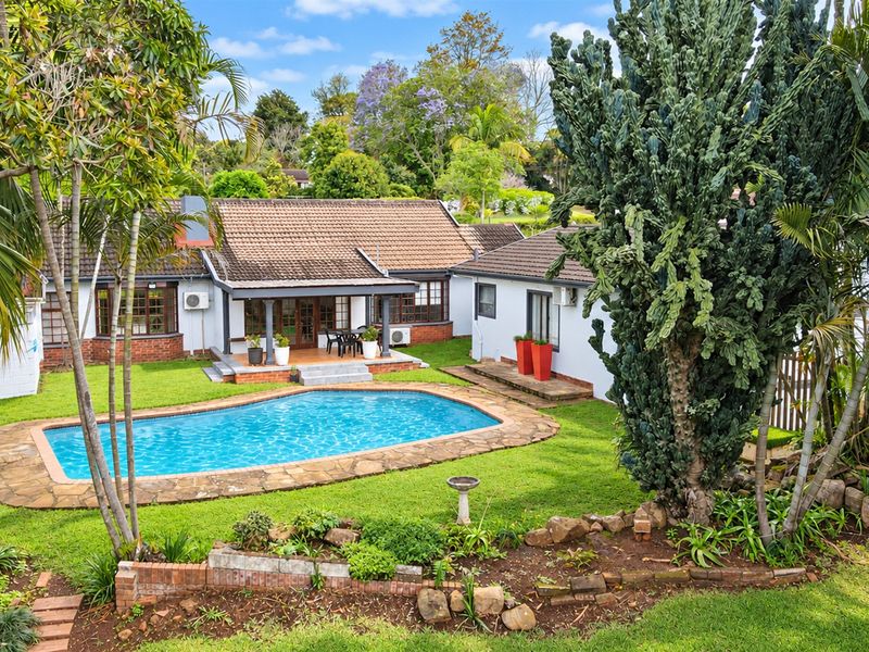 A well-maintained rear exterior view featuring a swimming pool with stone coping, a covered patio area, and established garden landscaping.