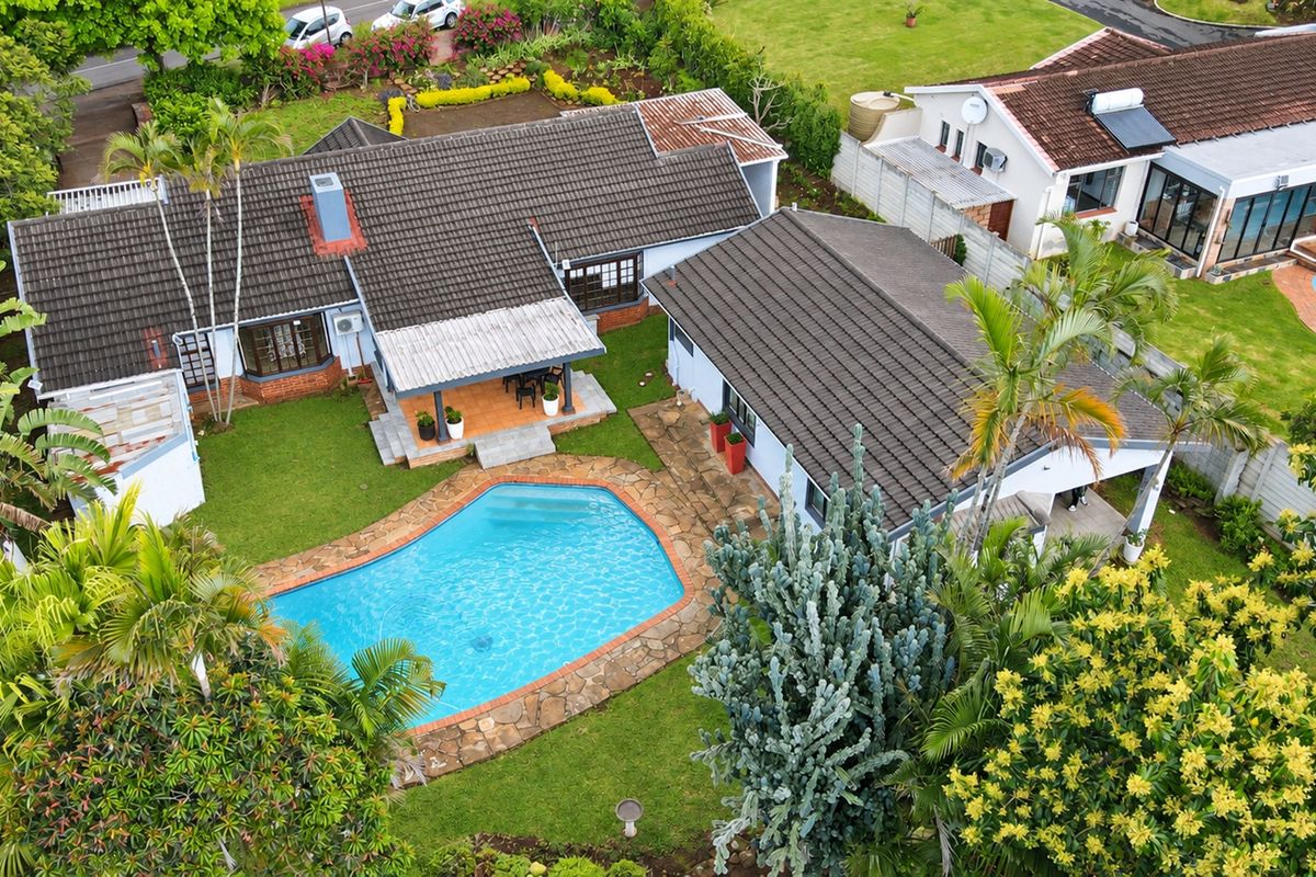 Aerial view of a residential property featuring a swimming pool, stone patio surround, and a tiled roof structure set within a landscaped garden.
