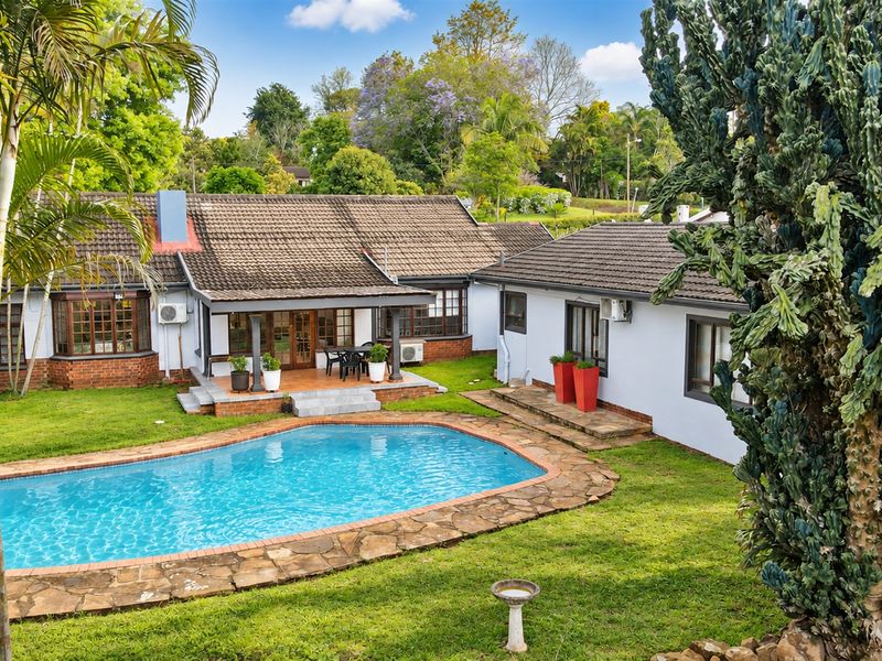 Rear view of the property featuring a swimming pool with stone coping, a covered patio area, and a well-maintained garden.