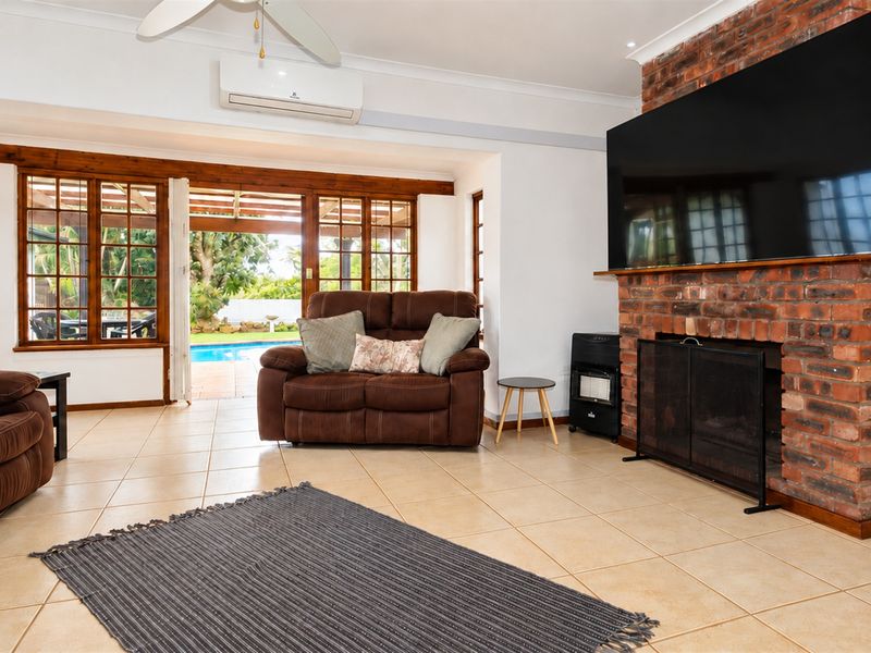Spacious living room featuring an exposed brick fireplace, ceramic tile flooring, and timber-framed sliding doors providing direct access to the outdoor pool area.