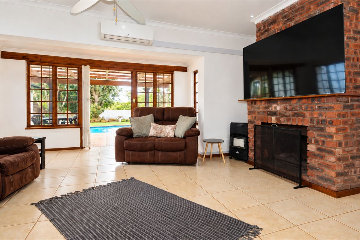 Spacious living room featuring an exposed brick fireplace, ceramic tile flooring, and timber-framed sliding doors providing direct access to the outdoor pool area.
