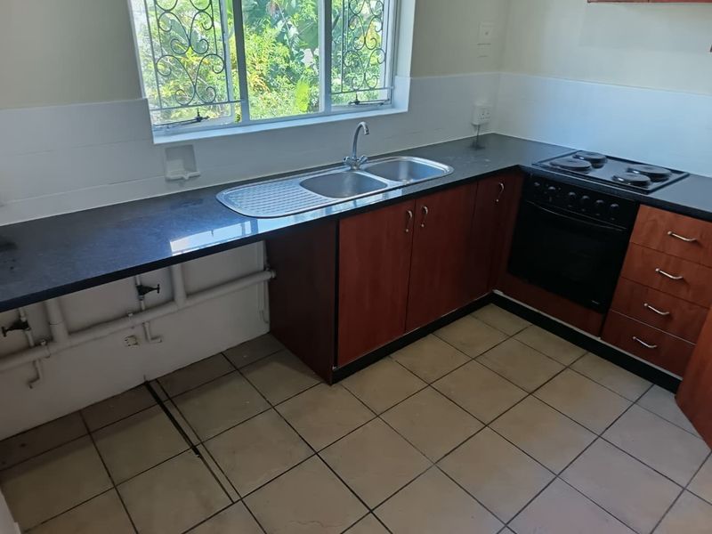 A functional kitchen featuring dark laminate cabinetry, a double sink, built-in oven, electric hob, and tiled flooring with a window overlooking the garden.