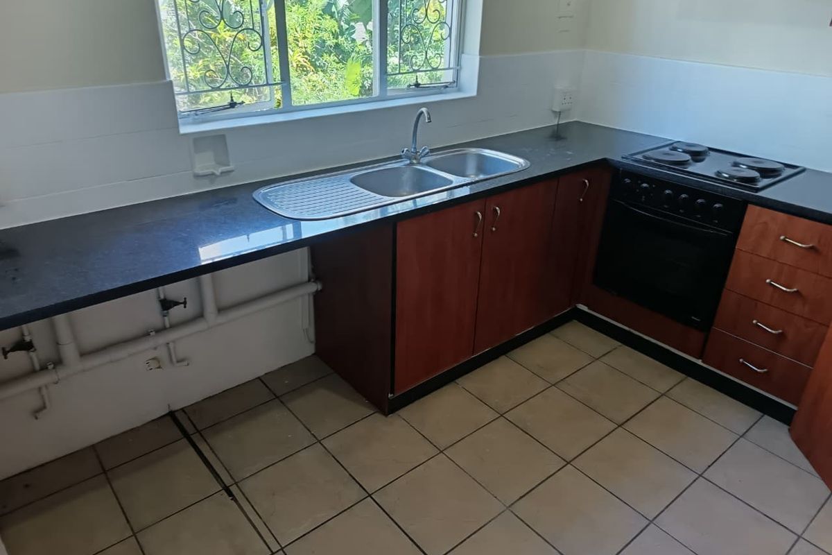 A functional kitchen featuring dark laminate cabinetry, a double sink, built-in oven, electric hob, and tiled flooring with a window overlooking the garden.