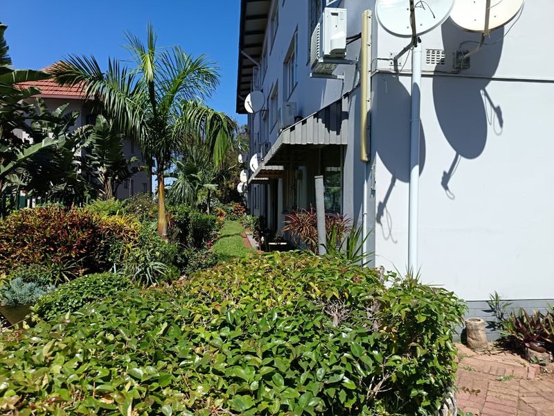 A view of the rear exterior of the flat building, featuring a landscaped garden area, paved pathways, and mounted satellite dishes.