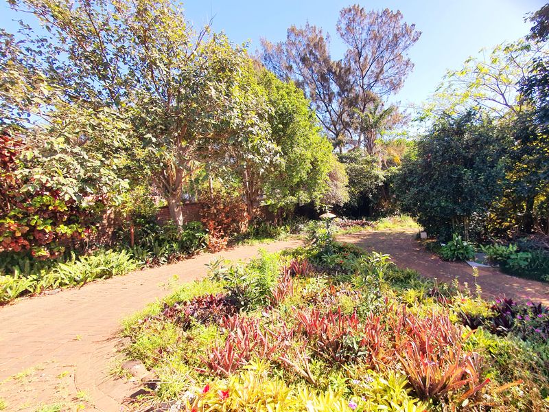 A lush, established garden featuring mature trees, diverse shrubbery, and a winding brick-paved pathway leading through the property grounds.