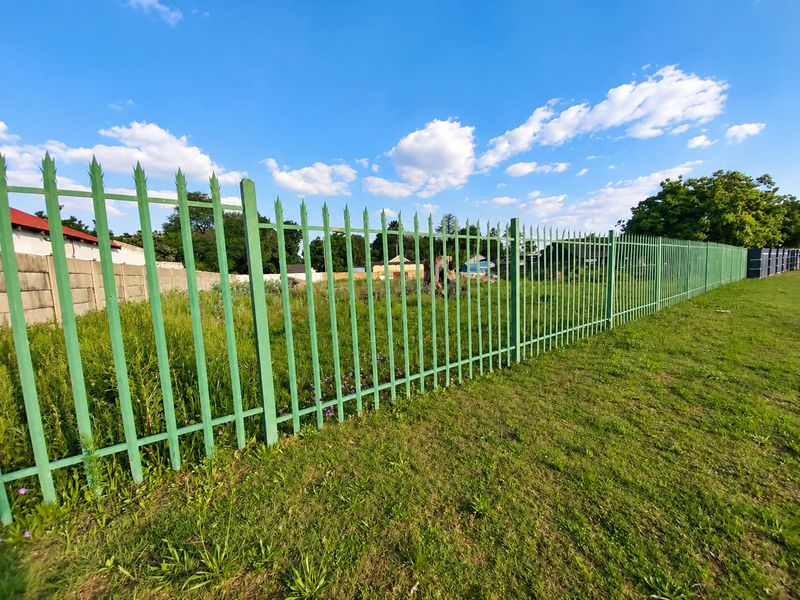 A vacant residential plot secured with green steel palisade fencing, featuring a grass verge and clear visibility of the undeveloped land.