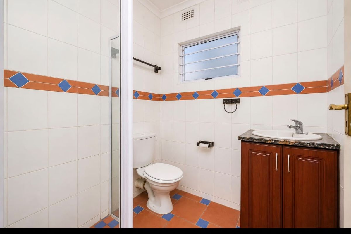 A functional bathroom featuring a vanity with granite countertop, a toilet, and a shower enclosure with dated decorative wall tiling.
