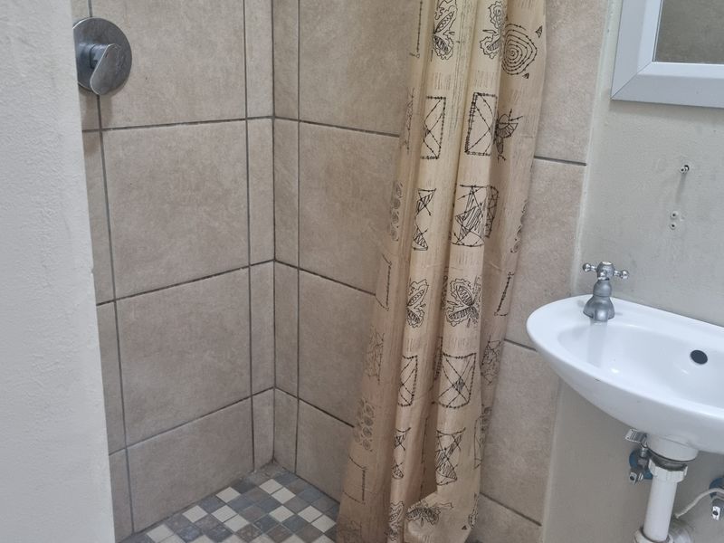 A compact bathroom featuring a tiled shower enclosure and a wall-mounted basin with exposed plumbing, requiring cosmetic updates.