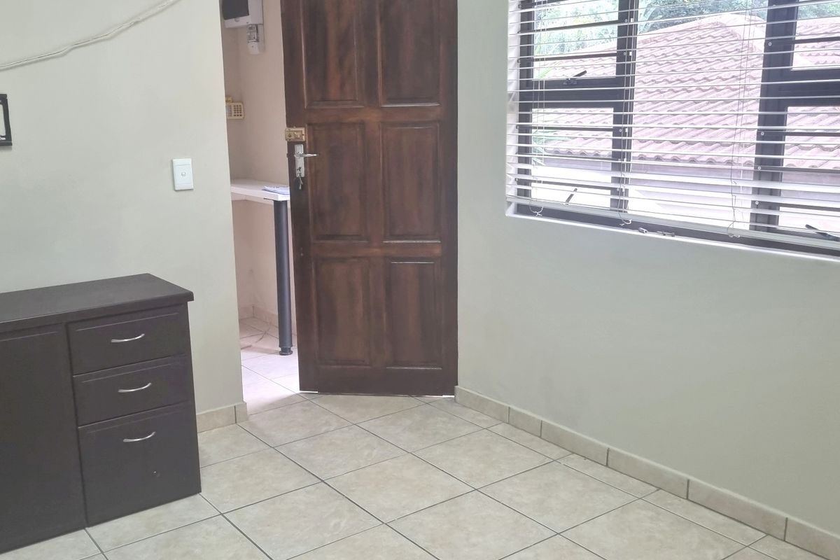 A simple bedroom featuring tiled flooring, a timber door, and venetian blinds, with an adjacent utility area visible through the doorway.
