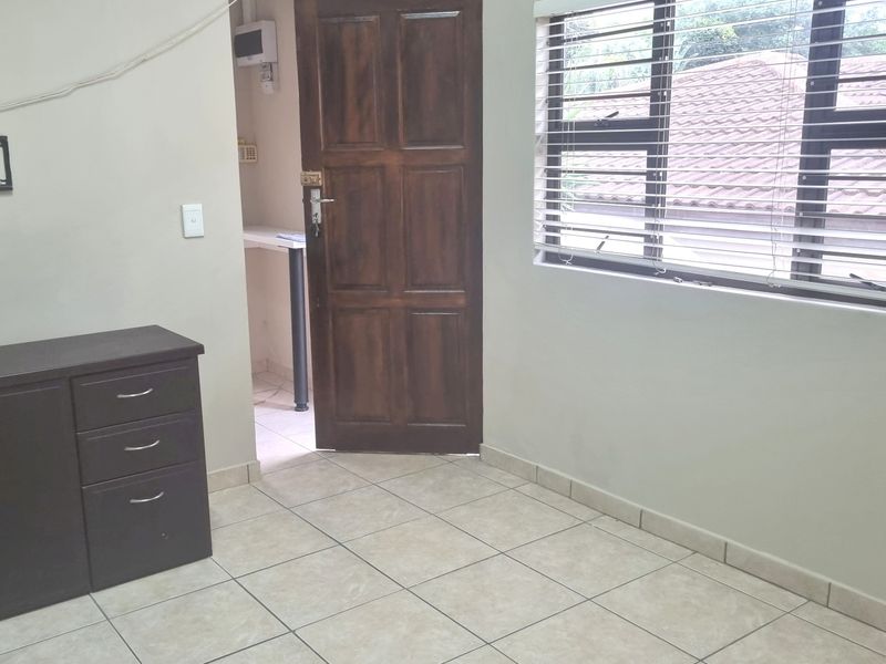 A simple bedroom featuring tiled flooring, a timber door, and venetian blinds, with an adjacent utility area visible through the doorway.