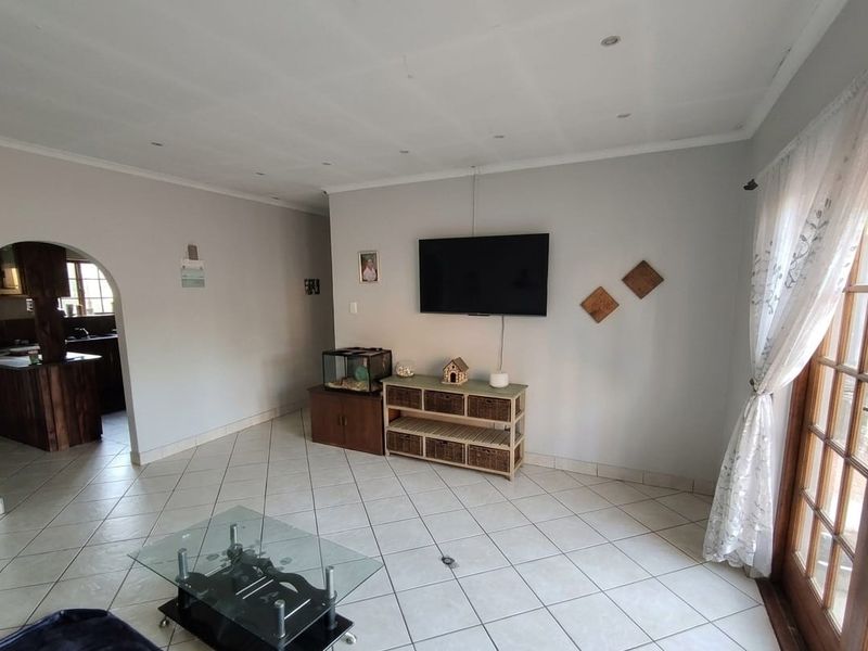 A living room featuring tiled flooring, recessed lighting, and an archway leading to the kitchen, with access to the outdoors via wooden framed glass doors.