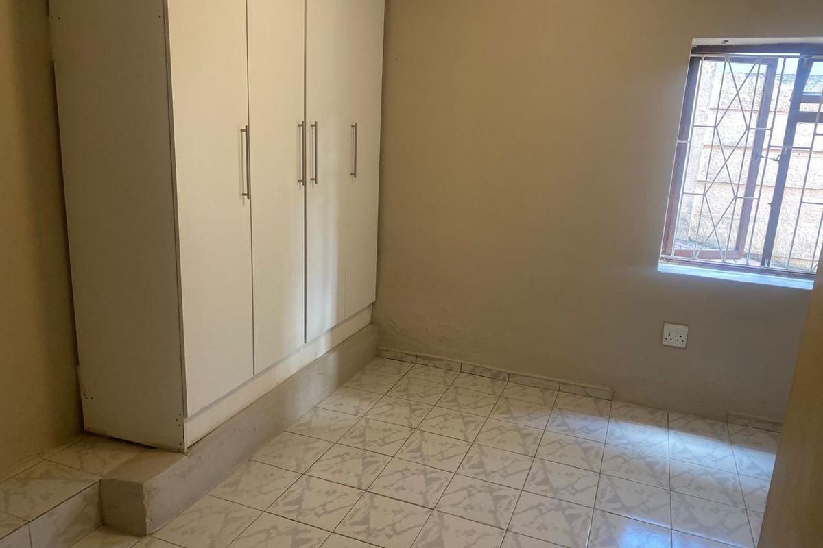 A compact bedroom featuring tiled flooring, a built-in wardrobe, and a window fitted with security bars.