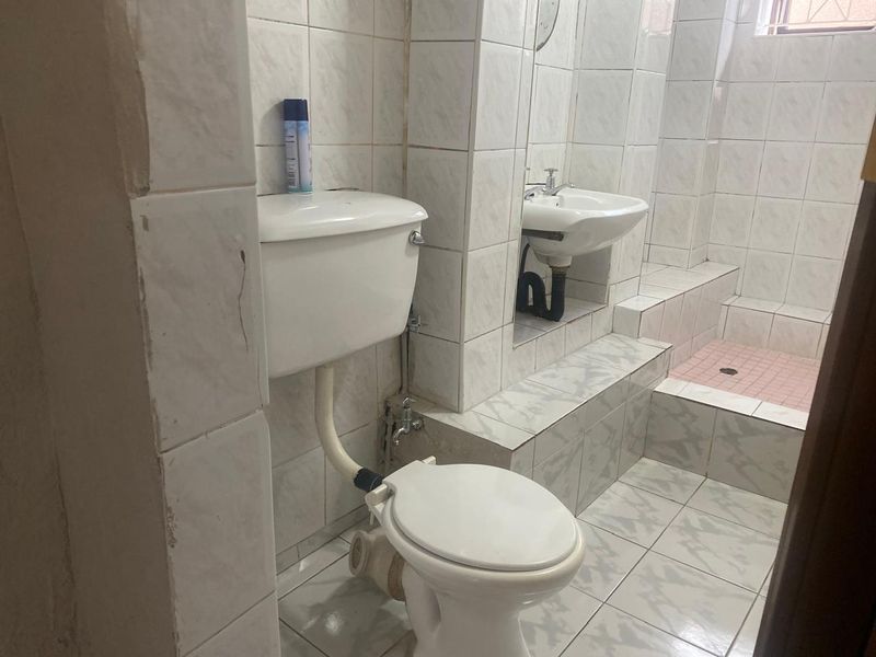 A functional bathroom featuring a toilet, pedestal basin, and a raised tiled shower area with dated ceramic finishes and exposed plumbing.