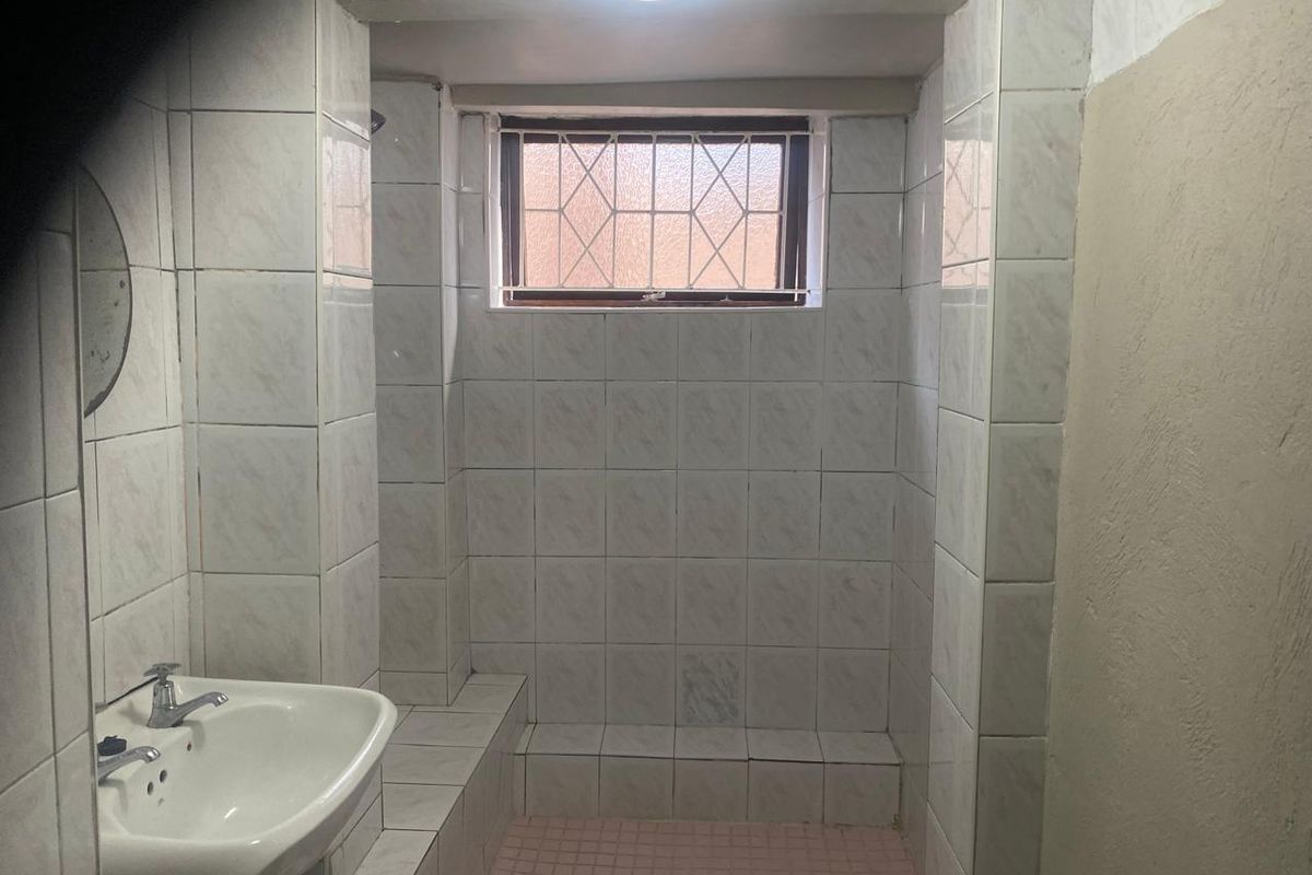A functional bathroom featuring a tiled shower enclosure, pedestal basin, and security-barred window, requiring cosmetic updates to improve its overall condition.