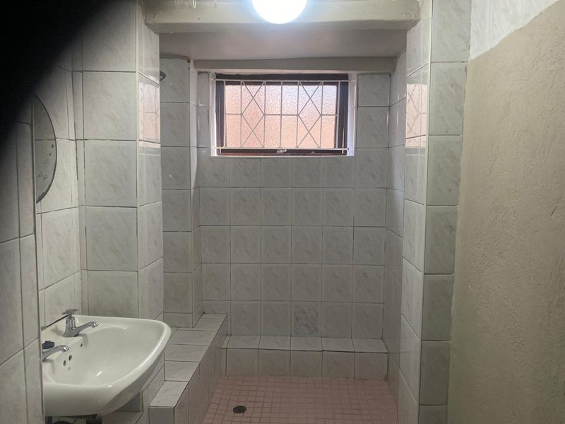A functional bathroom featuring a tiled shower enclosure, pedestal basin, and security-barred window, requiring cosmetic updates to improve its overall condition.