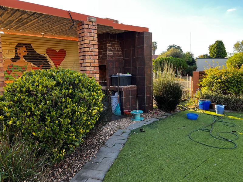 Outdoor patio area featuring a built-in brick braai, tiled surround, and corrugated metal wall cladding, overlooking a landscaped garden space.