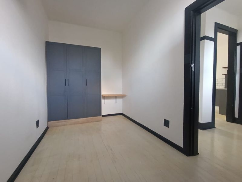 A bright, empty bedroom featuring a modern grey built-in wardrobe, a small wall-mounted shelf, and contrasting black door frames and skirting boards.
