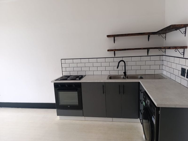 A modern, compact kitchen featuring dark grey cabinetry, subway tile backsplash, built-in oven, electric hob, and floating timber shelving.