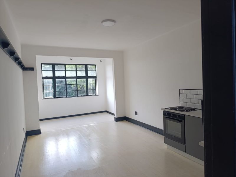 An open-plan living and kitchen area featuring a built-in oven, hob, subway tile backsplash, and large windows, presented in a clean, vacant state.