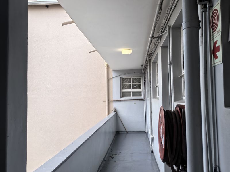 An exterior apartment balcony corridor featuring a fire hose reel, exposed utility piping, and a concrete floor finish.