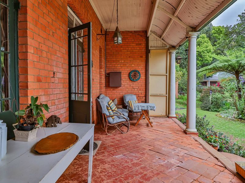 A charming covered veranda featuring exposed brick walls, terracotta flooring, and classical columns, overlooking a lush, established garden.