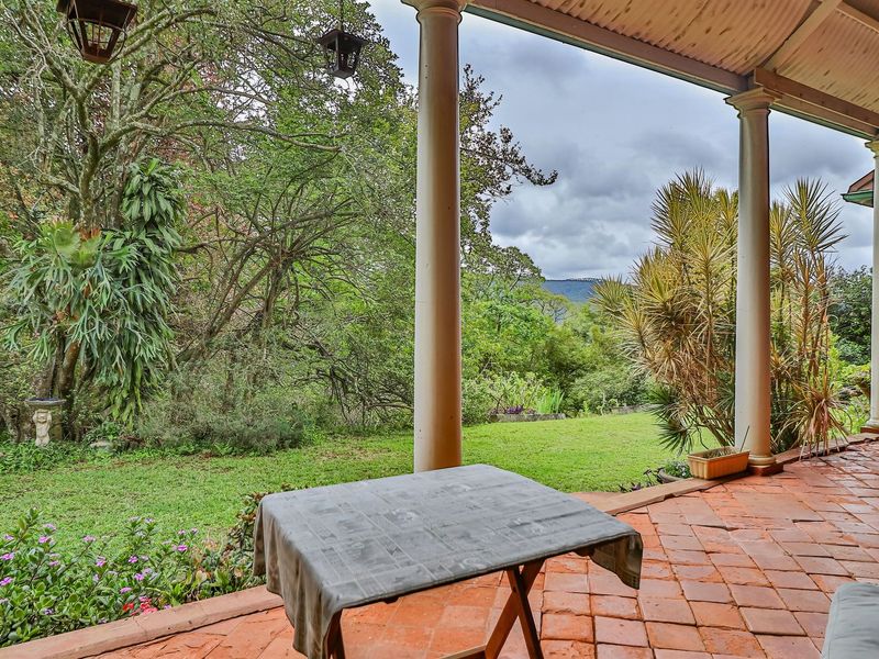 A covered outdoor patio featuring terracotta tiling and classical columns, overlooking a lush garden with mature trees and scenic views.