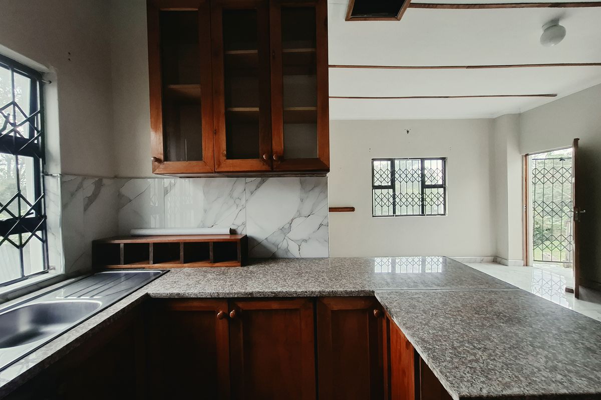 A functional kitchen featuring wooden cabinetry, granite countertops, and a marble-look splashback, with an open-plan layout leading to an adjacent living area.