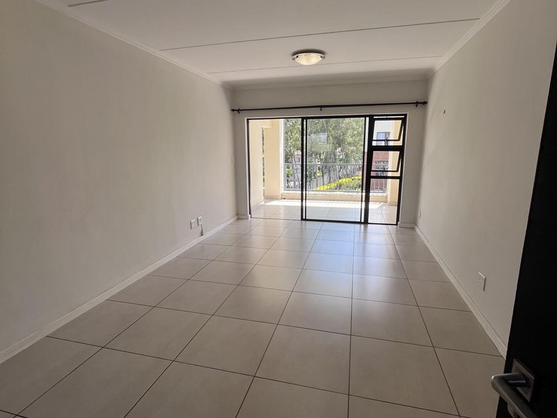 A bright, empty living room featuring neutral tiled flooring, coved ceilings, and a large sliding glass door leading to an outdoor balcony.