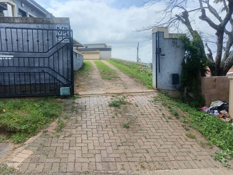 Front entrance view featuring a paved driveway, metal security gate, and boundary wall, showing signs of wear and maintenance requirements.