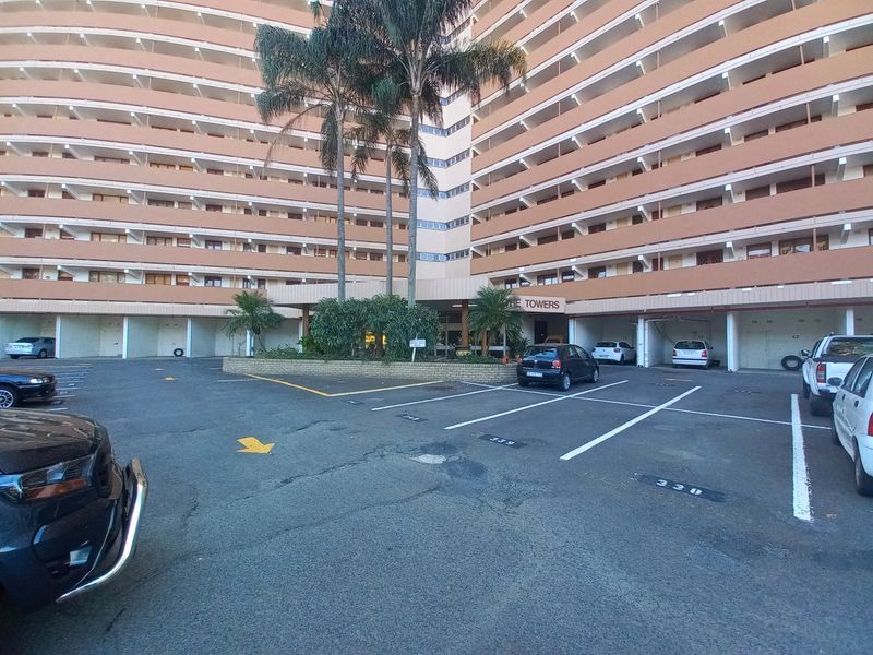 Exterior view of a multi-story apartment building featuring a paved parking area with designated bays and established palm trees at the entrance.