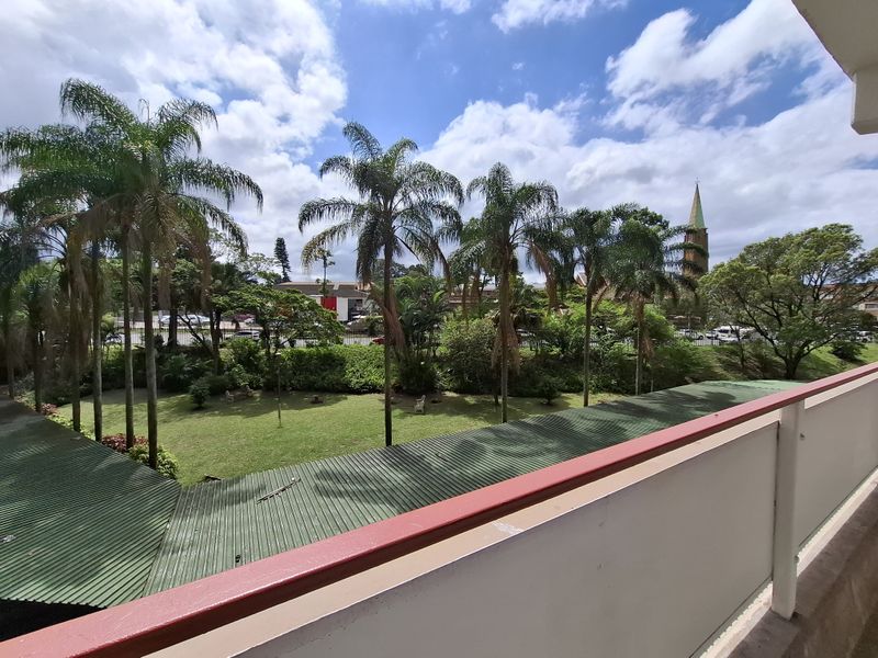 A view from an elevated balcony overlooking a landscaped garden area with palm trees and a distant church spire.