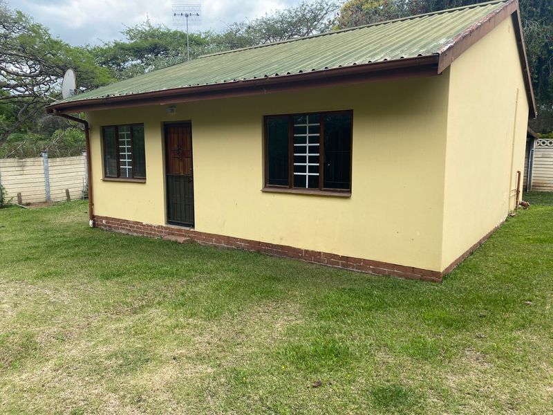 Rear view of a cottage featuring a corrugated metal roof, brick plinth, and security bars on windows, set within a grassed garden area.