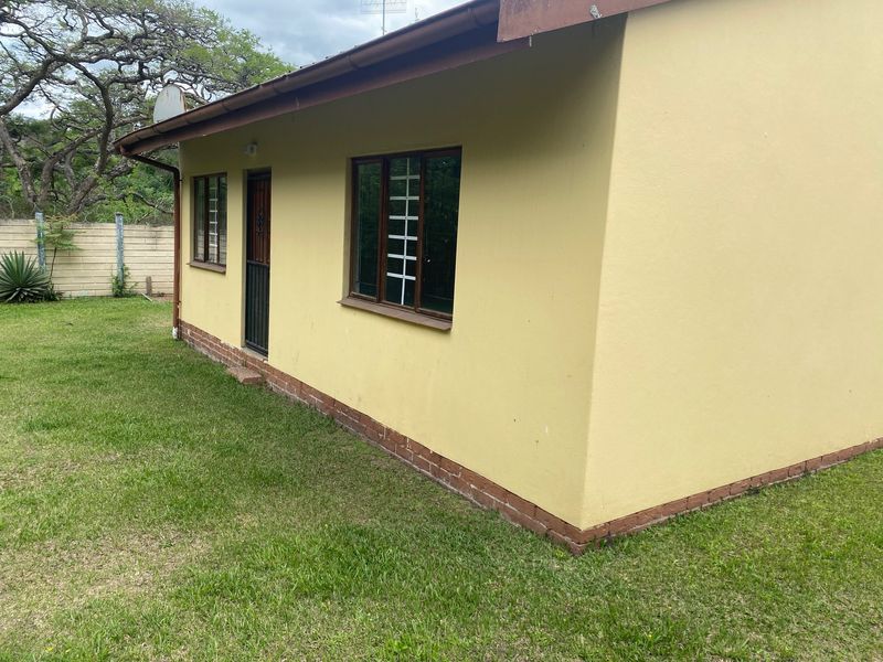 Rear view of the cottage featuring yellow rendered walls, a brick plinth, security-barred windows, and established garden surroundings.