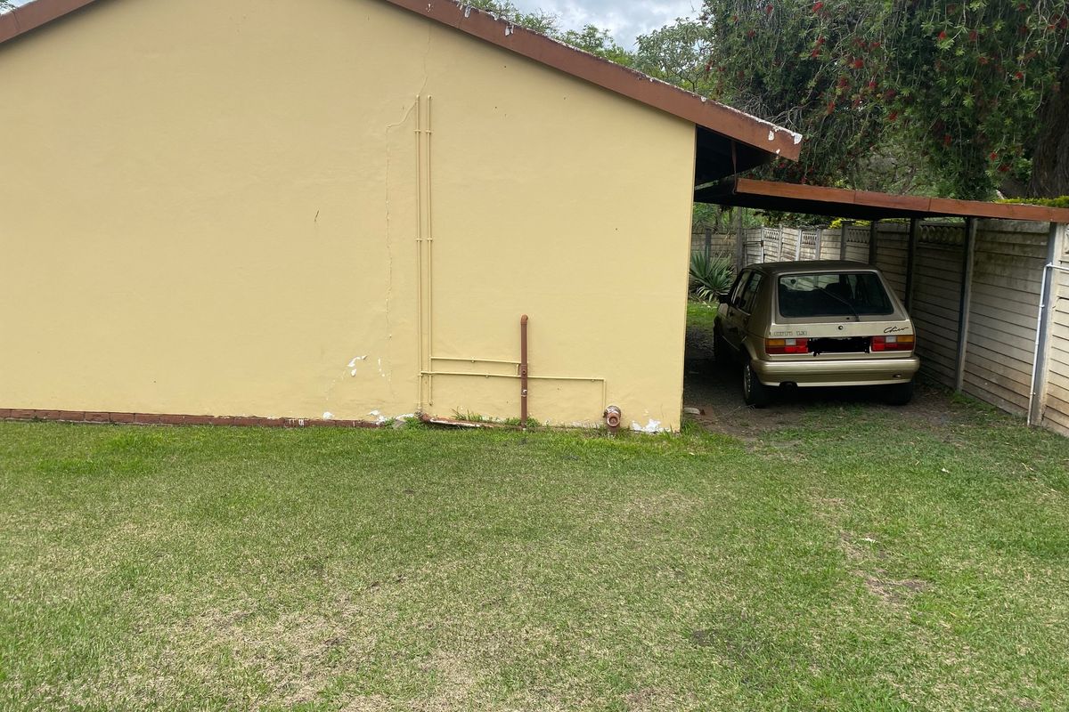 Rear exterior view of the cottage featuring a carport, lawn area, and precast boundary walling with visible signs of wear on the exterior plaster.