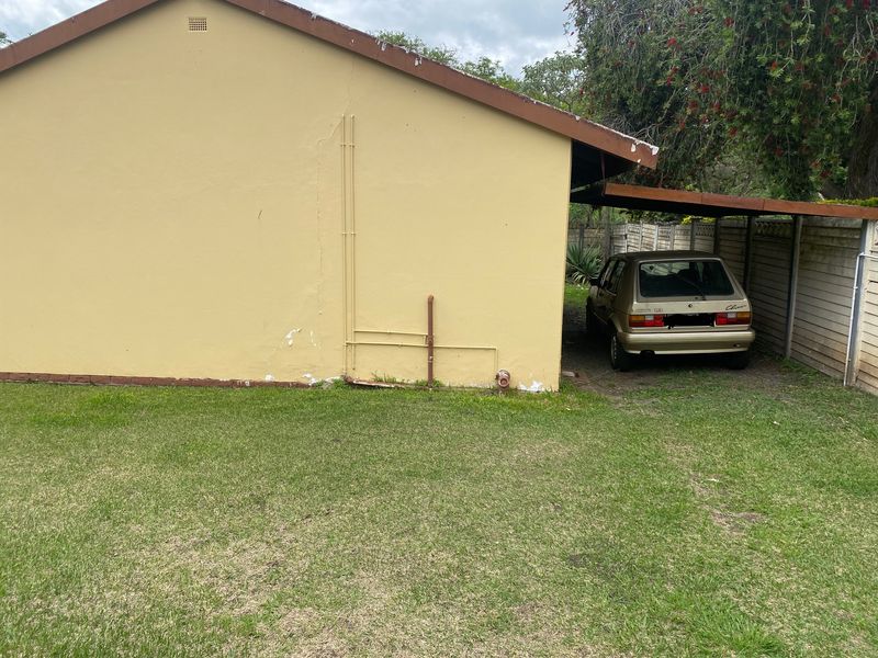 Rear exterior view of the cottage featuring a carport, lawn area, and precast boundary walling with visible signs of wear on the exterior plaster.
