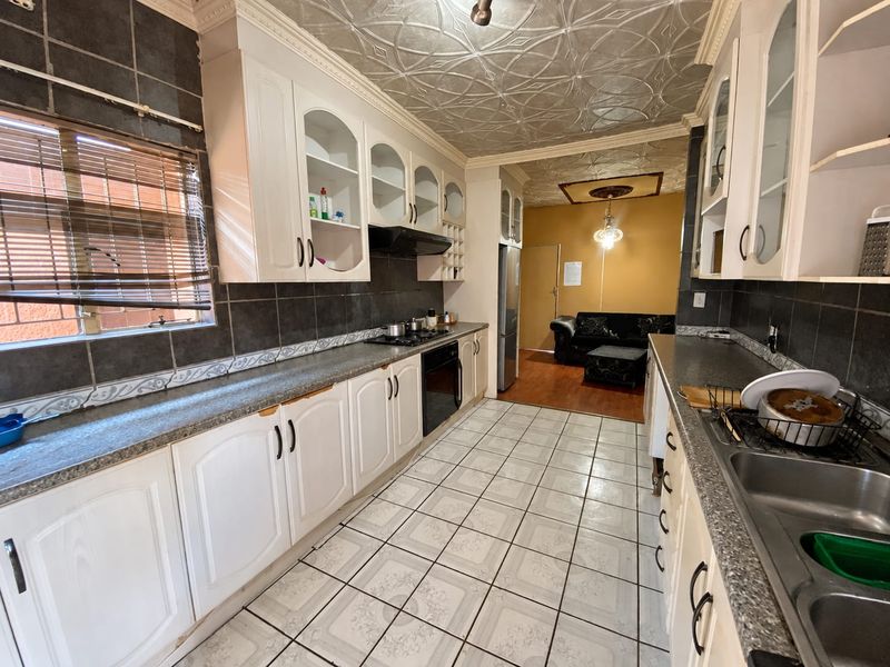 A functional kitchen featuring white cabinetry, laminate countertops, built-in oven and hob, and decorative ceiling tiles, leading into an adjacent living area.