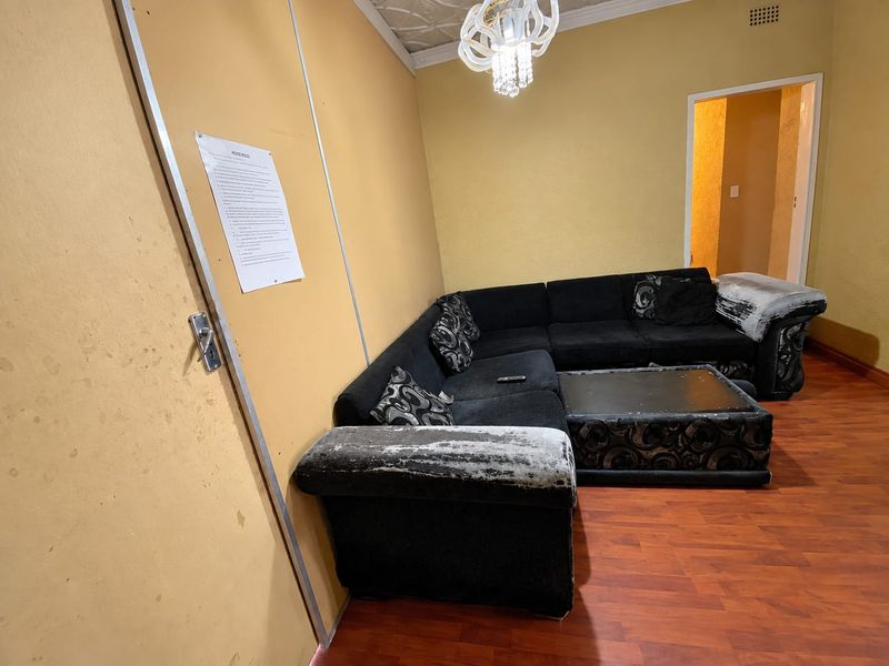 A living room featuring laminate flooring, decorative ceiling tiles, and a crystal chandelier, currently showing signs of wear on the wall surfaces.