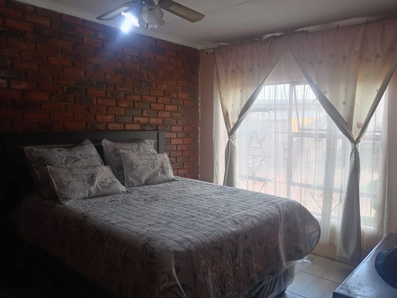 A bedroom featuring an exposed brick accent wall, tiled flooring, and a ceiling fan, with natural light entering through a window with security bars.