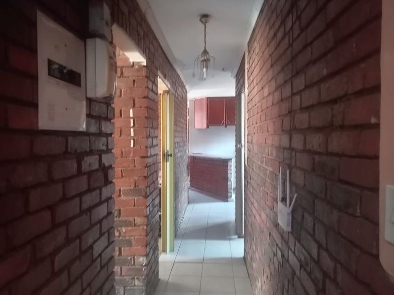 A narrow hallway featuring exposed brick walls and tiled flooring, leading towards an open-plan living area with kitchen cabinetry visible in the background.
