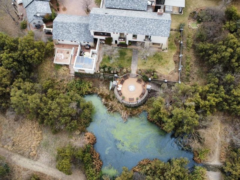 Aerial view of a large residence featuring a slate roof, solar panels, and a stone-walled circular patio overlooking a natural pond.