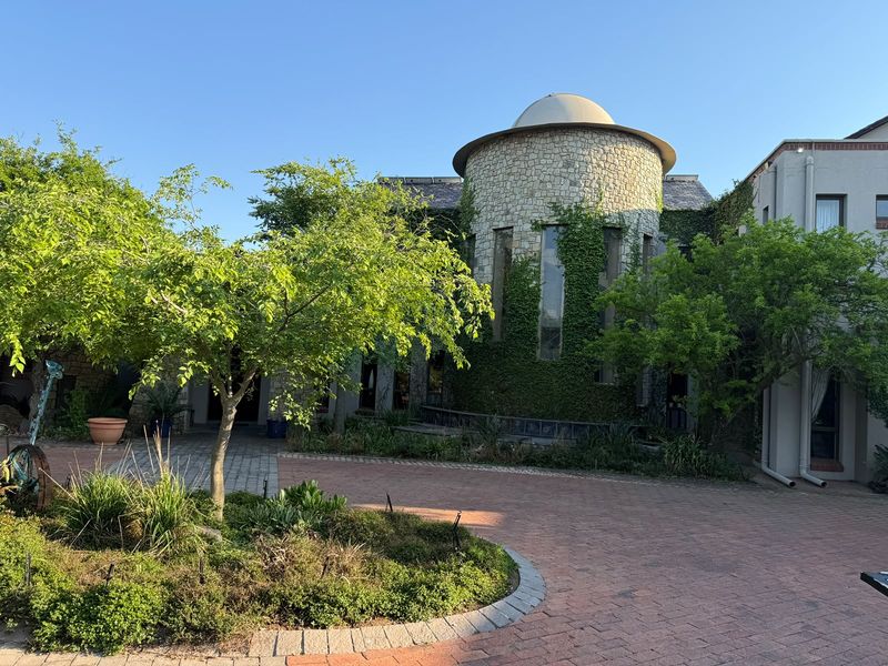 A unique architectural front facade featuring a stone turret, domed roof, and a well-maintained brick paved driveway with established garden landscaping.