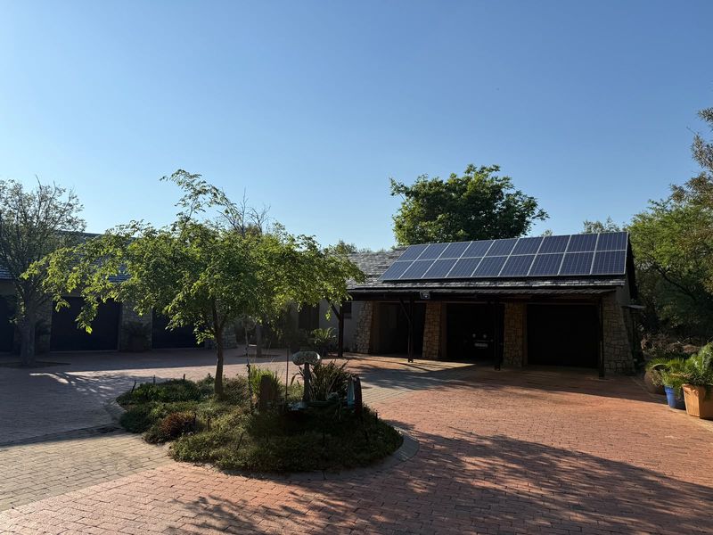 A spacious front exterior featuring a paved driveway, stone-clad garage structure, and roof-mounted solar panels set within a landscaped garden area.