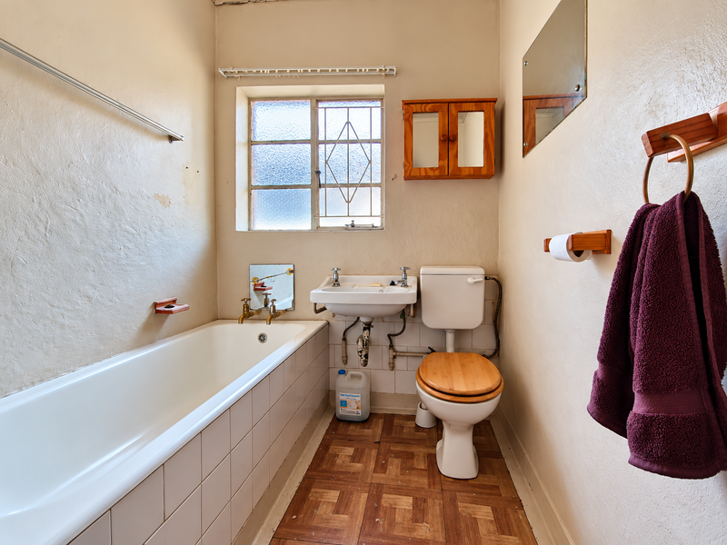 A dated bathroom featuring a standard bathtub, wall-mounted basin, and toilet, with original tiled finishes and wood-look flooring in need of modernization.