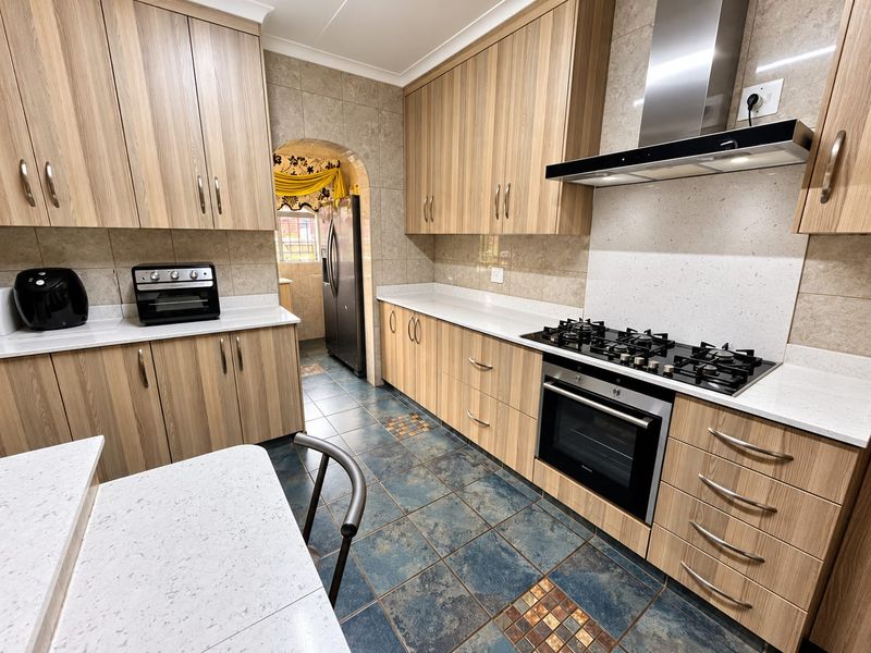 A well-maintained kitchen featuring wood-finish cabinetry, stone countertops, a gas hob with extractor fan, and tiled flooring, with an adjacent scullery area.
