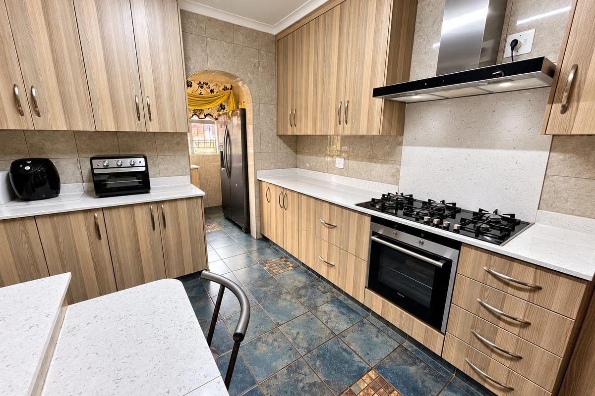 A well-maintained kitchen featuring wood-finish cabinetry, stone countertops, a gas hob with extractor fan, and tiled flooring, with an adjacent scullery area.