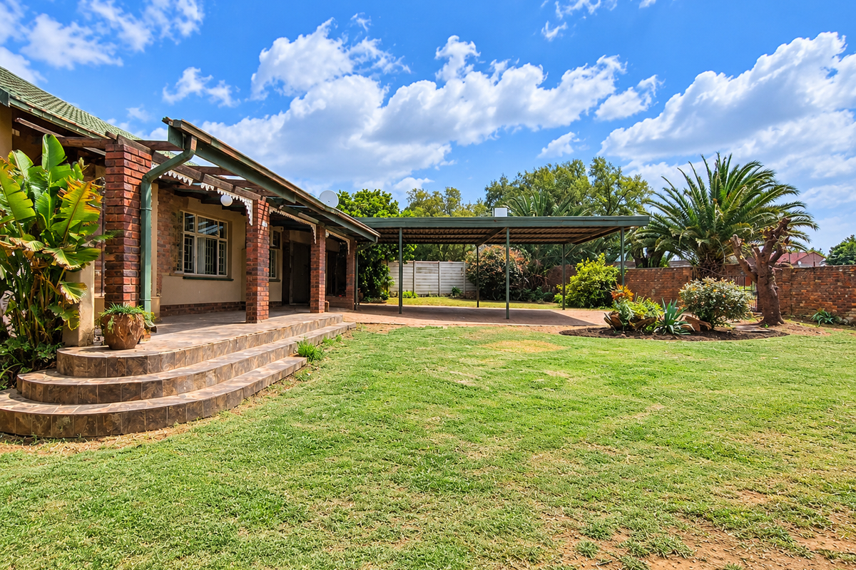 Spacious rear garden featuring a covered patio with brick pillars, a large carport, and established landscaping with a brick boundary wall.
