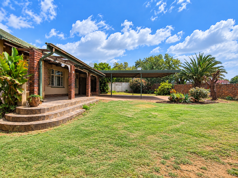 Spacious rear garden featuring a covered patio with brick pillars, a large carport, and established landscaping with a brick boundary wall.