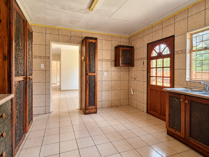 A spacious, empty kitchen featuring tiled walls and floors, built-in wooden cabinetry, and direct access to the exterior through a timber door.