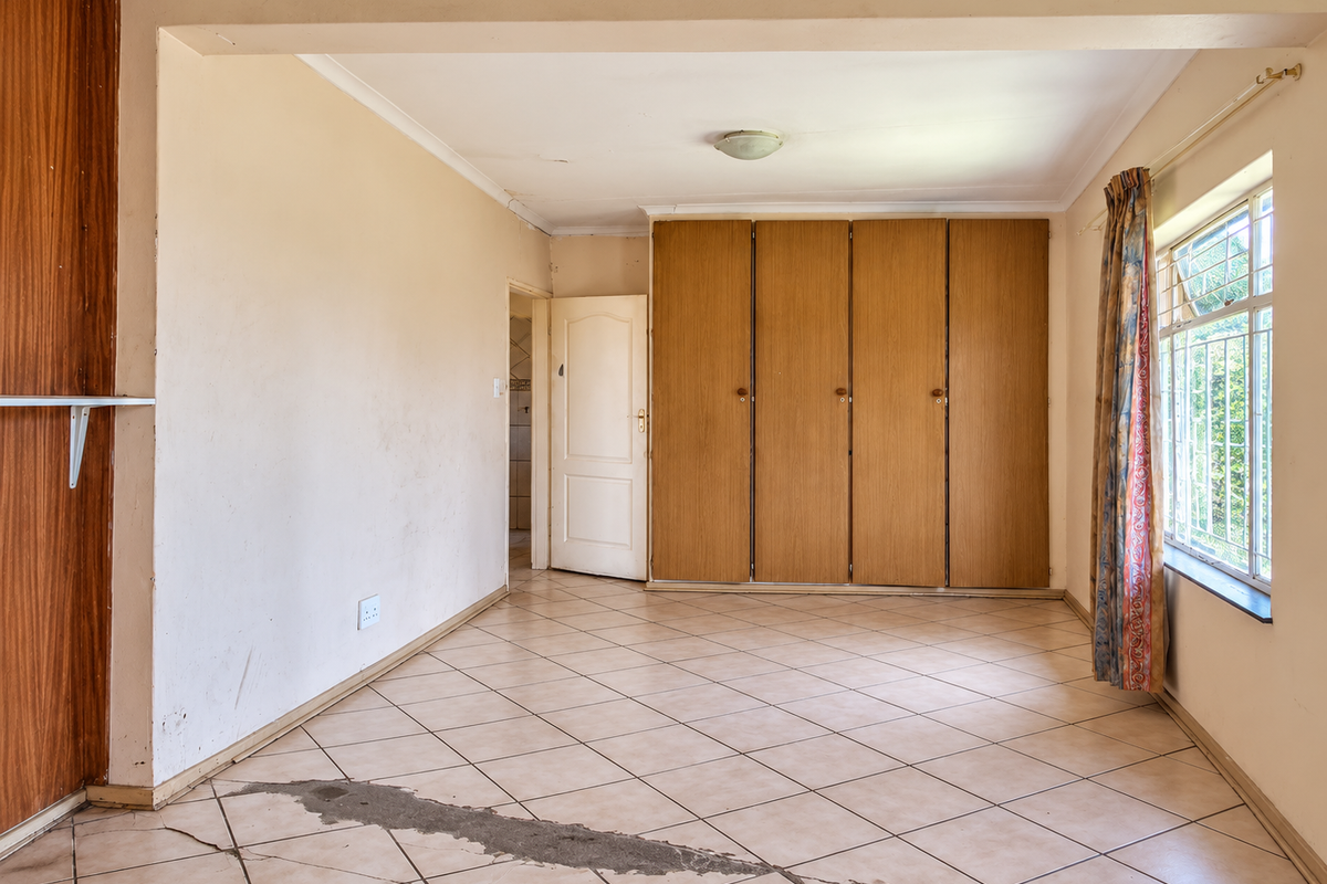 A vacant bedroom featuring built-in wooden cupboards, tiled flooring, and security bars on the window. The room requires cosmetic updates and floor repairs.